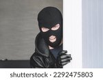 Small photo of Stealthy criminal wearing black balaclava sneaking into house through window or glass door. copy space