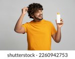 Small photo of Smiling curly haired Indian man holding bottle with hair shampoo, looking at cosmetic product isolated on gray background, studio shot. Hair care concept