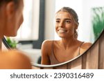 Smiling beautiful confident woman with perfect makeup wearing tank top looking in mirror standing at home. Natural beauty, skin care, morning routine concept