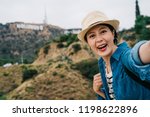 elegant tourist smiling lovely with the Hollywood sign in the background. happy tourist girl taking self-portrait picture with smartphone during summer vacation in famous landmark in US.