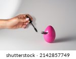 Woman’s hand holding eye mascara to apply on lashes on pink Easter egg. Make up and beauty Easter concept. Creative minimal Easter composition.                               