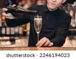 Cropped photo of young bartender pouring champagne into a glass behind bar counter in bar, closeup view, alcohol bottles in background