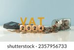 Small photo of VAT in 2024 concept. VAT wooden letter and 2024 number on wooden block and glass coin jar with calculator. Value Added Tax.