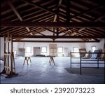 The image presents a spacious artist's studio with exposed wooden beams, white walls, and multiple canvases and art materials scattered throughout, suggesting a creative and work-intensive environment