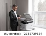 Adult successful man in a gray business suit standing near a copier with a cup of coffee