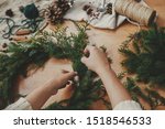 Hands holding fir branches, and pine cones, thread, berries, scissors on wooden table. Christmas wreath workshop. Authentic stylish still life. Making rustic Christmas wreath.
