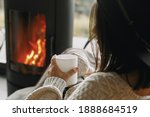 Stylish woman in knitted sweater relaxing with warm cup of tea at modern black fireplace with view on mountains. Cozy warm moments at cold season. Young female resting