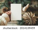 Christmas card mock up. Hand holding empty greeting card on background of christmas paper stars, wooden tree, pine branches and cones on rustic wood.  Space for text. Seasons greetings template