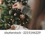 Hands decorating christmas tree with stylish bauble in atmospheric festive room. Merry Christmas! Winter holidays preparation. Woman in cozy sweater putting vintage bell on tree