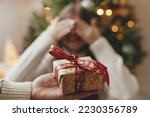 Merry Christmas and Happy Holidays! Hand holding christmas gift box on background of happy woman closing eyes waiting for surprise. Exchanging presents at christmas tree in atmospheric room
