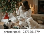Cozy christmas morning. Beautiful woman in pajamas opening stylish christmas gift with cute white dog and cat at fireplace and decorated tree in festive living room. Merry Christmas!