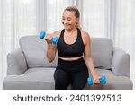 Small photo of Athletic and sporty senior woman engaging in body workout routine, sitting on sofa and lifting dumbbell at home as concept of healthy fit body with body weight lifestyle after retirement. Clout