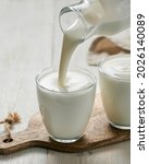 Small photo of Pouring kefir, buttermilk or yogurt with probiotics. Yogurt flowing from glass bottle on white wooden background. Probiotic cold fermented dairy milk drink. Vertical