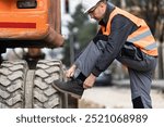 A construction worker in safety gear is kneeling to tie his boot next to large machinery on a bustling city street, preparing for the day