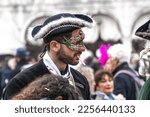 Small photo of Venice, Italy - 02.19.2022: Handsome young man in carnival mask wearing tricorn with an unlit cigarette in his mouth at Venice carnival at the street in Venice, Italy