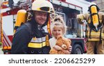 Small photo of Firefighter hug rescued little girl with teddy bear. Frightened child rejoices in rescue. At background firefighters after extinguishing fire next to fire truck. Concept of saving lives, fire safety