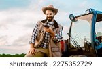 Small photo of Cheerful young male driver at farm standing at tractor, tapping and texting on smartphone and resting. Farming concept. Happy man farmer having rest and using mobile phone for chatting. Outside.