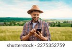 Small photo of Caucasian handsome man in hat standing in grassland and using mobile phone. Outside. Good-looking young male farmer tapping and texting on smartphone. Gadget for farming. Chatting while working.