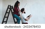 Small photo of Caucasian pretty woman standing on the ladder in the room measuring wall with a measure tape and her husband coming to her with a tablet devise as showing some ideas of renovation on it.