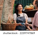 Happy Asian woman in an apron receiving payment on the outdoor market. Vendor with a card machine smiling and looking at the customer.