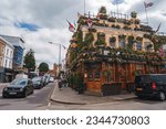 Small photo of London, UK. July 10, 2023. Churchill Arms covered with flowers and leaves. Cars moving on road under cloudy sky. People walking on footpath by famous pub of London.