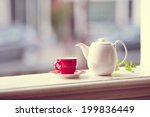 Red cup of tea with white teapot on wooden windowsill with blurred city in a background.  Vintage look.