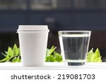 Disposable coffee cup and glass of water on windowsill with city in background.