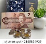 Small photo of Saguenay, Canada, june 2022 : close up of old discontinued canadian 2 dollars bill in paper, canadian passport in back ground with blue vase and a mini plant, some money in the front