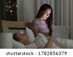 Woman housewife sitting on bed in the night bedtime and using laptop notebook computer connect to internet while husband sleeping beside her. Idea for social addict lifestyle of modern couple.