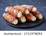 Hungarian dessert kurtos kalacs or chimney cake filled with cream sprinkled with icing sugar on top served on a black plate on a dark concrete background, horizontal orientation, close-up