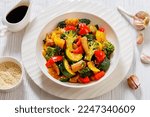vegetable stir-fry of bell pepper, onion, zucchini, baby corn in cobs, broccoli florets poured with sticky soy sauce in white bowl on white wood table, horizontal view