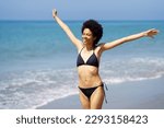 Carefree smiling African American female traveler in swimwear strolling with raised arms on coast along sea in tropical resort on summer day and enjoying freedom