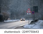 Small photo of Cars driving down a wet mountain road in winter with a slippery road and ice warning sign covered in snow and ice and snow on the sides of the road and on trees