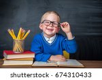 Happy cute clever boy is sitting at a desk in a glasses with raising hand. Child is ready to answer with a blackboard on a background. Ready for school. Back to school. Apple and books on desk