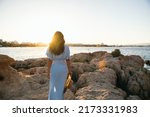 Back view of unrecognizable young woman traveler with long hair in stylish maxi dress with straw hat in hand standing on massive rocky boulders. She is admiring sea against cloudless sunset sky 