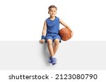 Small photo of Smiling boy in a jersey holding a basketball and sitting on a blank panel isolated on white background
