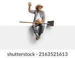 Small photo of Full length portrait of a mature farmer with a spade sitting on a blank panel and waving isolated on white background