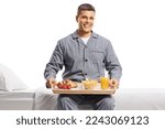 Smiling young man in pajamas sitting on a bed with a breakfast tray isolated on white background