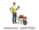 Construction worker holding a brick and standing behind a wheelbarrow with pile of bricks isolated on white background