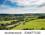 Rolling fields and trees of lush green British countryside stretch to the horizon under wispy clouds and blue sky