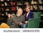 Small photo of Senior bearded gray-haired grandfather with his grandson enjoy memories watching family photo album sitting on armchair and floor at home in living room with an authentic aristocratic interior.