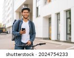 Medium shot of smiling handsome male courier with thermo backpack standing with bicycle in city street and using navigation app on phone. Delivery man looking for client address looking smartphone.