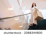 Small photo of Low-angle view of smiling young woman shopper holding on escalator handrail and riding escalator going down in shopping mall, looking away, paper bags with purchases in hands, blurred background.