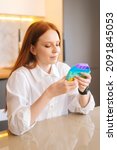 Small photo of Vertical portrait of attractive redhead young woman playing fidget toy new trendy silicone toy anti stress pop it and simple dimple, sitting at table in kitchen room with modern light interior.