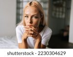 Close up face of thoughtful blonde female sitting alone in living room and serious looking at camera holding hands on chin, thinking over health problems, feeling sadness, boredom, apathy.