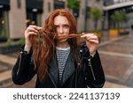 Small photo of Portrait of despaired young woman touching wet hair after autumn rain standing on beautiful city street, looking at camera. Front view of worried female untangling hair after being caught in downpour.