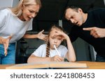 Small photo of Portrait of angry young parents yelling and scolding together lazy little daughter sitting at table, doing homework, sad looking at camera. Concept of parent disciplining child for bad education.