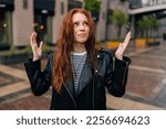 Portrait of frustrated red-haired young woman touching wet hair after autumn rain standing on beautiful city street. Front view of upset female untangling hair after being caught in rain.