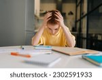Small photo of Portrait of exhausted pupil boy tired from studying holding head head with hands sitting at desk with paper copybook, looking down. Frustrated child schoolboy doing homework at home.