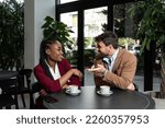 Small photo of Young man and African American woman sitting in cafeteria drinking coffee flirting talking laughing and seducing outside working hours and colleague in restaurant away from busy street